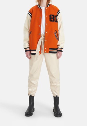 Le modèle porte une veste universitaire orange à manches crème, un haut blanc, un pantalon crème à cordon de serrage et des bottes noires.