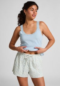 Woman with curly hair wearing a light blue lace-trimmed tank top and white floral shorts, adjusting her top with both hands.