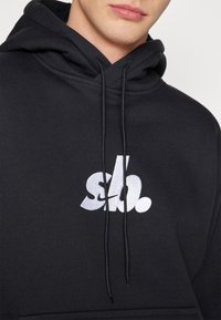 Czarna bluza z kapturem uszyta z mieszanki bawełny, z przednią kieszenią, sznurkiem do regulacji kaptura oraz dużym białym haftowanym logo na piersi.