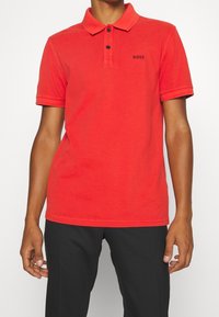 Man med en klar röd kortärmad polo skjorta med "BOSS"-logotyp på bröstet, kombinerad med svarta byxor, mot en vit bakgrund.