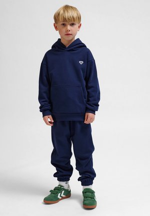 Granatowy hoodie z przednią kieszenią i logo, zestawiony z pasującymi dresami. Zielone zamszowe trampki z rzepami. Czysty, swobodny design.
