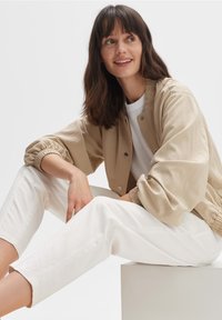 Beige bomberjack met elastische manchetten en een drukknoopsluiting aan de voorkant, gecombineerd met witte cropped broeken. Gladde stof en een losse pasvorm.