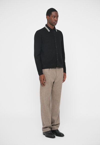 Mies seisoo, päällään musta napillinen villatakki, jossa on valkoiset raidat kauluksessa, väljiä beigejä housuja ja mustia kenkiä yksinkertaista valkoista taustaa vasten.