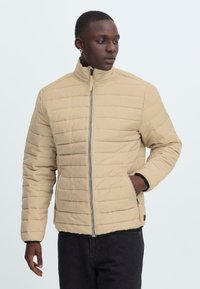 En ung person står iført en beige pufferjakke, hænderne i lommerne, mod en lys baggrund, hvilket udstråler en afslappet, moderne stil.