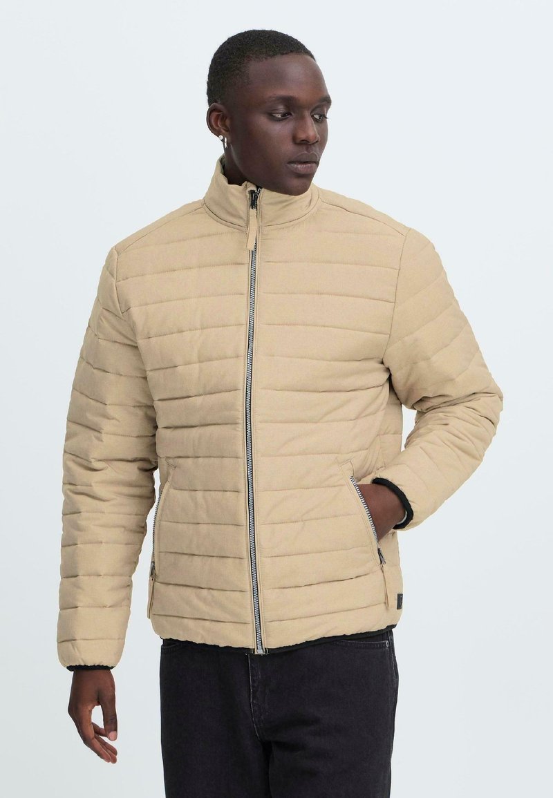 En ung person står iført en beige pufferjakke, hænderne i lommerne, mod en lys baggrund, hvilket udstråler en afslappet, moderne stil.