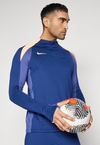 Tengerészi hosszúujjú sportfelső narancssárga részletekkel, cipzáras gallérral és hüvelykujjlyukakkal, fekete-fehér mintás focival párosítva.