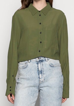 Femme portant une chemise boutonnée manches longues de couleur vert olive rentrée dans un jean taille haute délavé clair, debout devant un fond uni.