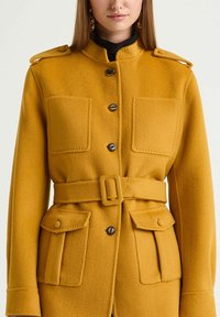 Manteau en laine jaune avec un col montant, une fermeture boutonnée à l'avant, une taille ceinturée et quatre poches à rabat. Finition avec des accents métalliques sur les épaules.