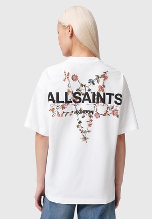 Lyshåret kvinde vendt væk iført en hvid skjorte med blomsterdesign og teksten "ALLSAINTS LONDON" på ryggen.