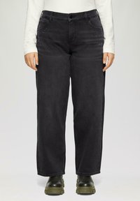Schwarze Jeans aus Denim mit lockerer Passform, verziert mit dekorativen, metallischen Nieten in der Nähe der Taschen. Getragen mit einem weißen Langarmshirt und schwarzen Stiefeln.