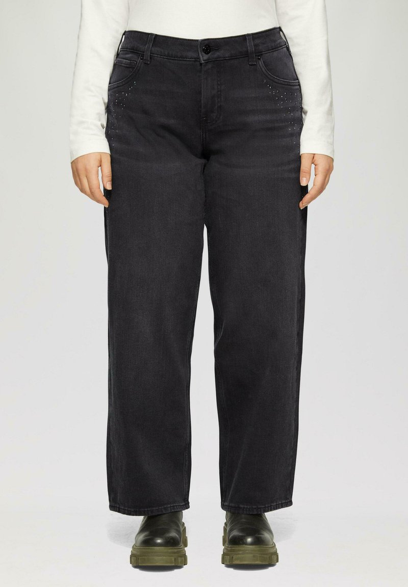 Schwarze Jeans aus Denim mit lockerer Passform, verziert mit dekorativen, metallischen Nieten in der Nähe der Taschen. Getragen mit einem weißen Langarmshirt und schwarzen Stiefeln.