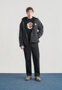 Μαύρο μπουφάν με φερμουάρ και επένδυση από συνθετική γούνα, μαύρο μπλουζάκι με στρογγυλό γραφικό, μαύρο παντελόνι και μπεζ παπούτσια με υφή στη σόλα πάνω σε μπλε δάπεδο.