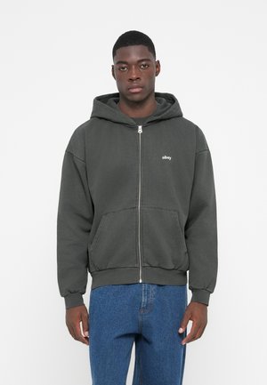 Zip-up hoodie in dark gray fabric with a kangaroo pocket, ribbed cuffs, and a white "obey" logo. Model wears blue jeans.