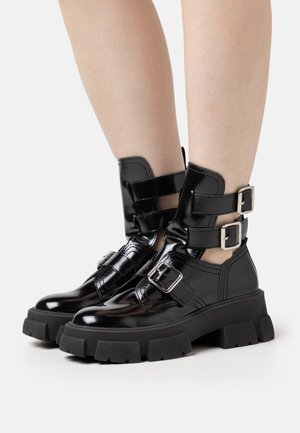 Bottines en cuir verni noir avec semelles épaisses, présentant deux sangles ajustables à boucle et une finition texturée sur la partie supérieure.