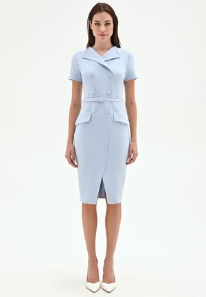 Femme debout, portant une robe bleu clair ceinturée, à double boutonnage, longueur genou, manches courtes et talons blancs pointus.