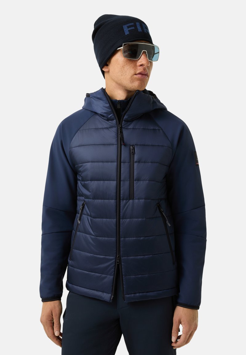 Marineblaue Steppjacke mit gesteppten Abschnitten, Reißverschlusstaschen und Kapuze. Kombiniert mit einer marineblauen Mütze und Sonnenbrille für den Außeneinsatz.