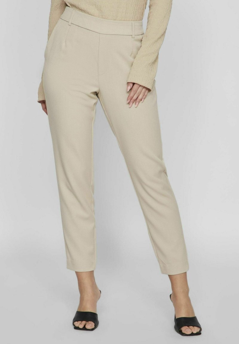Femme portant un pantalon beige taille haute sur mesure, un haut beige à manches longues et des mules à talons ouvertes noires, debout devant un fond uni.