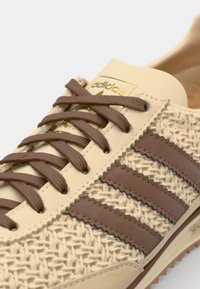 Baskets beige avec texture tissée, accents en cuir marron et trois rayures. Présente des lacets marron assortis et un logo doré sur la languette.