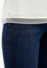 Witte top met een gestructureerde afwerking en een geschulpte kanten rand, gelaagd over donkerblauwe denim jeans met een soepele,licht vervaagde textuur.