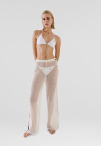 White bikini top with halter neck; sheer, knitted white pants with black side stripes; model standing with arms crossed.