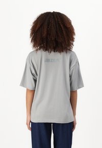 Šedé oversized tričko s krátkým rukávem, hladký povrch a jemné logo potisknuté na zadní straně. Nosené s tmavě modrými, volně střiženými kalhotami.