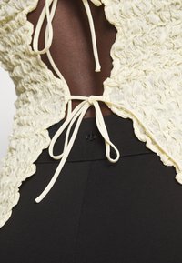 Krémový, texturovaný top s volánkovým lemem, se šňůrkami vzadu, v kombinaci s černými přiléhavými kalhotami. Design zdůrazňuje tvar a detail.