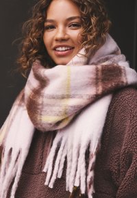 Rutig halsduk i rosa och brunt med fransiga ändar, gjord av mjukt tyg, virad runt halsen och kompletterad med en brun fleeceöverdel.