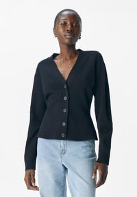 Kvinna i svart knäppt cardigan och ljusblå jeans, stående mot en enkel vit bakgrund.