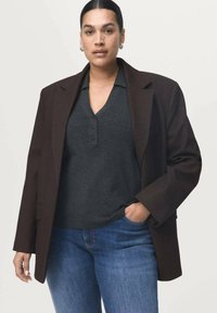 Blazer oversized marrón oscuro sobre un polo gris con botones, combinado con jeans azules. El blazer tiene hombros estructurados y una textura de tela suave.