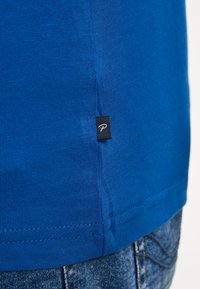 Modrá bavlněná tričko s žebrovanou strukturou a malou černou visačkou s bílým logem na lemu. Viditelné švy podél okrajů.