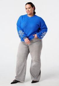 Femme aux cheveux foncés portant un pull bleu avec des motifs en sequins argentés sur les manches, un jean gris à jambes larges, et des chaussures noires pointues, debout.