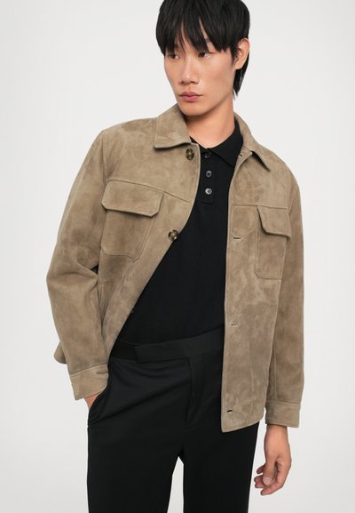 Suede jacket in light brown with a collared design, two chest pockets, and a button closure. Worn over a black polo and tailored pants.