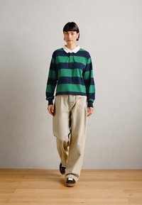 Groen en marineblauw gestreepte polo met lange mouwen en een witte kraag, gecombineerd met beige hoge broeken en bruine sneakers.