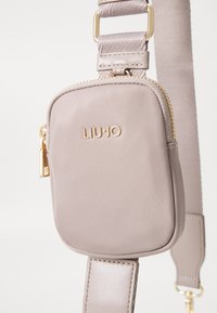 Borsa a tracolla beige chiaro con una texture morbida, hardware dorato e chiusura con cerniera. Presenta un logo del marchio impresso sul davanti.