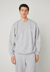 Felpa grigia realizzata in tessuto strutturato, con colletto rotondo e spalle scese. Piccolo etichetta verde sul petto, abbinata a pantaloni coordinati.