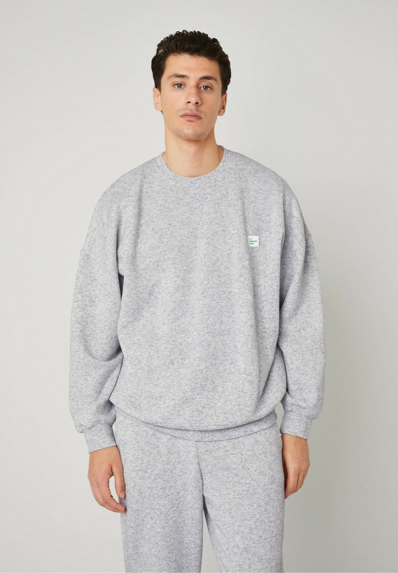 Felpa grigia realizzata in tessuto strutturato, con colletto rotondo e spalle scese. Piccolo etichetta verde sul petto, abbinata a pantaloni coordinati.