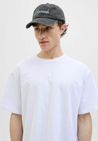 Sort bomuldskeppet med broderet "Originals" tekst i lyseblå. Båret med en ensfarvet hvid kortærmet T-shirt, modellen vendt let mod siden.