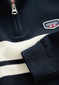 Marineblauer Strickpullover mit weißen Streifen, geripptem Bund, Halb-Reißverschluss und einem Superdry-Logopatch auf der Brust.