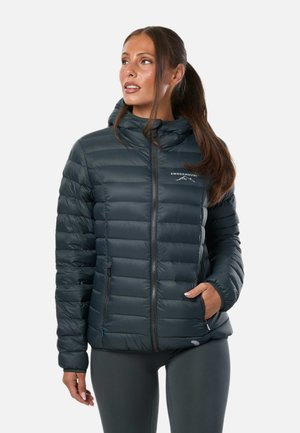Femme aux longs cheveux bruns attachés en queue de cheval, portant une veste matelassée foncée avec capuche et poches zippées, regardant vers sa gauche.