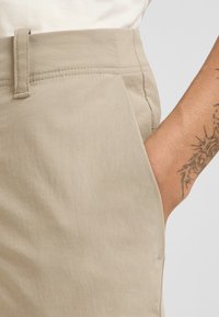 Pantaloni corti beige con una texture liscia, dotati di tasca laterale e passanti per cintura. Dettaglio di cucitura visibile lungo la vita.
