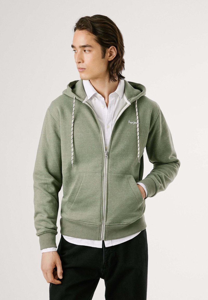 Sudadera verde con cremallera y cordones ajustables, bolsillos frontales y puños acanalados. Llevada sobre una camisa blanca con cuello y pantalones negros.