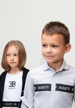 Zwei Kinder tragen Kleidung mit Bugatti-Marke. Der Junge hat ein hellgraues T-Shirt mit einem schwarzen Logo-Band. Das Mädchen trägt ein weißes T-Shirt und eine schwarze Jacke.