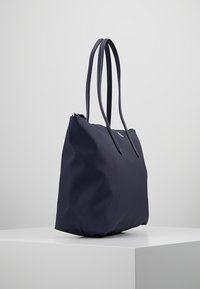 Sac cabas bleu marine en matériau texturé avec deux longues anses et une fermeture zippée. Présente un détail de logo subtil à l'avant.