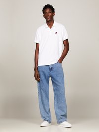 Camisa polo blanca con un pequeño logotipo de color en el pecho, combinada con jeans vaqueros ligeros de corte holgado y zapatillas blancas. Fondo neutro.