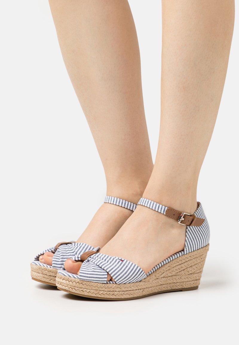 Jambes portant des sandales compensées à rayures bleues et blanches avec des brides à la cheville et des semelles en corde tressée sur un fond clair uni.