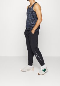 Dark blue patterned tank top, black joggers with zippers and white accents, and neutral sneakers with green highlights. Slim fit, athletic design.