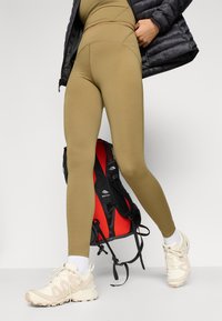 Khaški leggingi z visokim pasom, v paru s svetlimi supergami. Črna in rdeča nahrbtnik leži v bližini. Teksturovan pufferjakna.