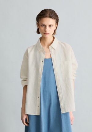 Junge Person mit kurzen Haaren trägt ein off-white Knopfhemd über einem lockeren blauen Kleid und steht vor einem einfarbigen hellgrauen Hintergrund.