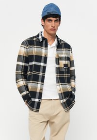 Camicia flanella a scacchi nera, beige e grigia con chiusura a bottoni e taschino sul petto; indossata sopra una maglietta polo bianca; abbinata a pantaloni beige.