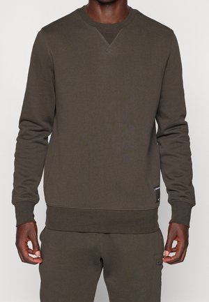 Mężczyzna w ciemnooliwkowej, długorękawowej bluzie i pasujących spodniach, stojący z ramionami luźno opuszczonymi przy bokach na jednolitym tle.